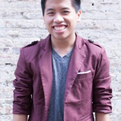 Frank pictured from the waist up smiling at the camera against a white brick wall wearing a maroon jacket and blue-grey t-shirt