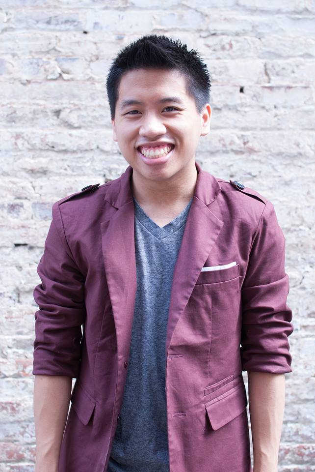 Frank pictured from the waist up smiling at the camera against a white brick wall wearing a maroon jacket and blue-grey t-shirt