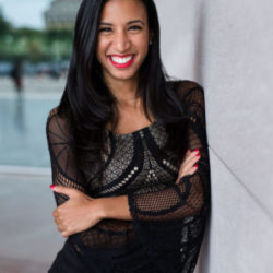 Kelsey, smiling with her arms folded wearing a black dress leaning against a white marble mall