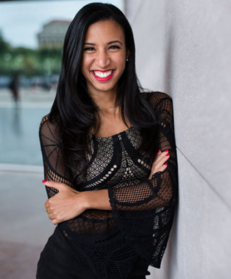 Kelsey, smiling with her arms folded wearing a black dress leaning against a white marble mall