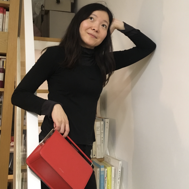 Shafei with her hand on her head and elbow leaning her against the wall, her other hand is holding a red bag and she is looking up away from the camera