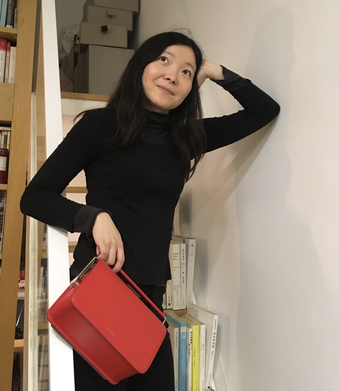 Shafei with her hand on her head and elbow leaning her against the wall, her other hand is holding a red bag and she is looking up away from the camera