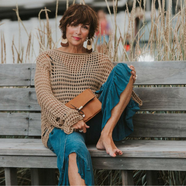 Debbie Love seated on a bench in a beach-like setting with one leg up, and holding a purse