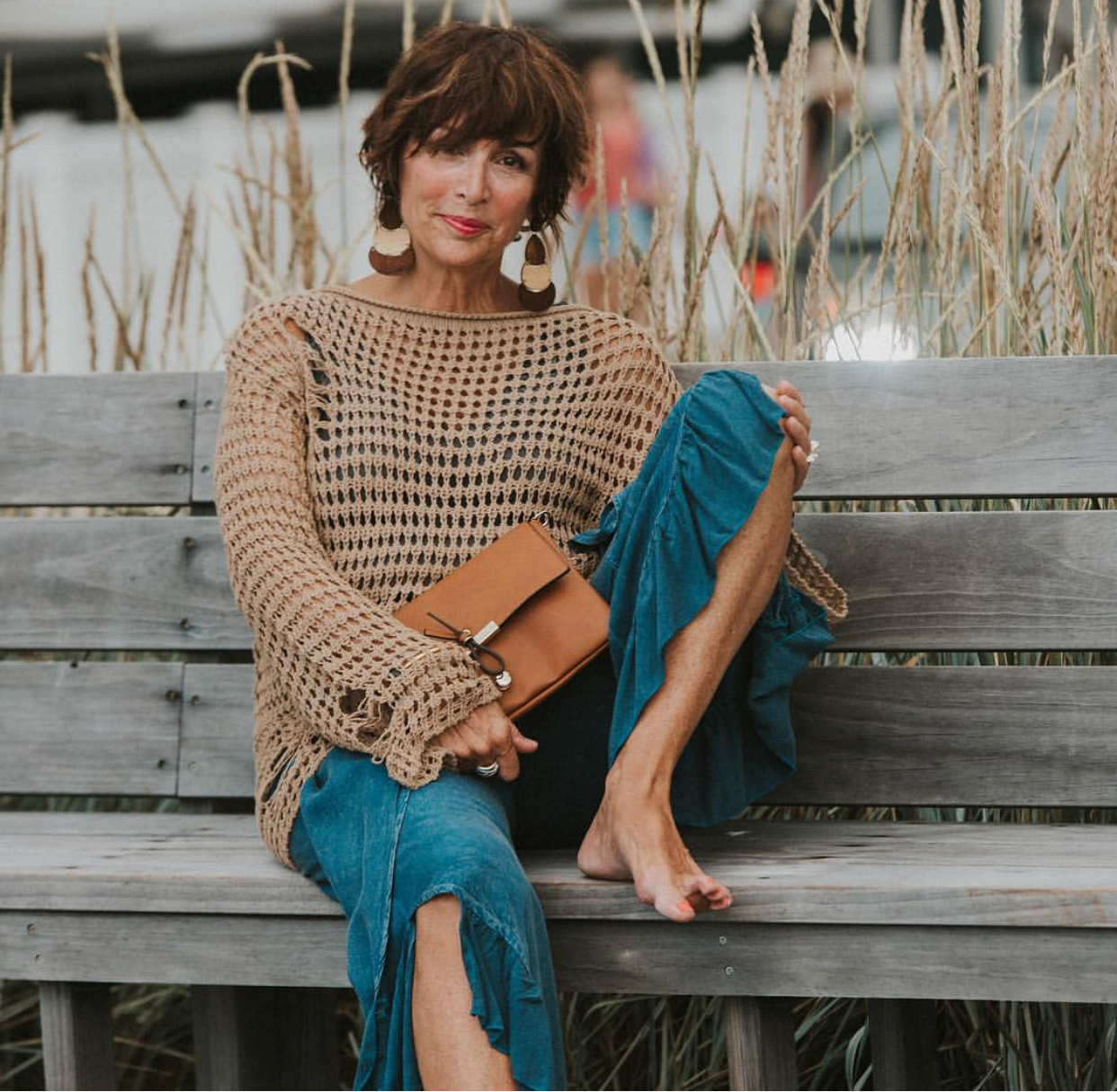 Debbie Love seated on a bench in a beach-like setting with one leg up, and holding a purse